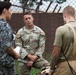 Three military personnel are engaged in a discussion outdoors near a chain-link fence. The central figure wears a US Air Force multicam uniform with the name tape 'MEANEY' and rank insignia visible. A person in what appears to be a foreign military uniform (blue digital camouflage) stands to the left, while a third individual in tan tactical gear with 'POLICE' visible on the back stands to the right.