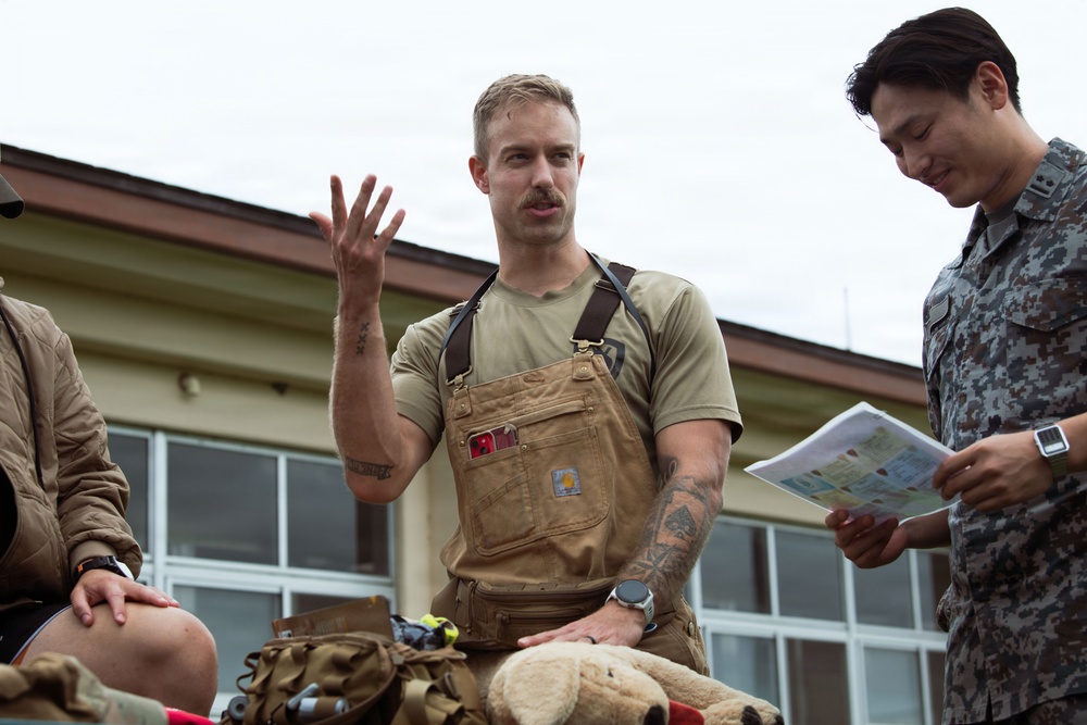 A man wearing Carhartt overalls and a tan t-shirt with visible arm tattoos is gesturing with his right hand, appearing to explain or brief something. He is seated or positioned near gear and a stuffed animal, while a person in blue digital camouflage military uniform reviews a document beside him. The setting appears to be an outdoor area near a building, suggesting a military or emergency services context.