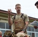 A man wearing Carhartt overalls and a tan t-shirt with visible arm tattoos is gesturing with his right hand, appearing to explain or brief something. He is seated or positioned near gear and a stuffed animal, while a person in blue digital camouflage military uniform reviews a document beside him. The setting appears to be an outdoor area near a building, suggesting a military or emergency services context.