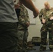A U.S. Air Force instructor is demonstrating how to don MOPP (Mission Oriented Protective Posture) gear, specifically chemical/biological protective overgarments, in what appears to be an indoor training environment. Two uniformed personnel in OCP (Operational Camouflage Pattern) are observing the demonstration, while a civilian or off-duty individual is partially visible in the foreground. The setting appears to be a classroom or briefing room.