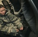 A female soldier wearing US Army ACU (Army Combat Uniform) digital camouflage pattern is seated in what appears to be an indoor office or administrative space. She appears to be putting on or adjusting a military boot near a large tire or vehicle component. The setting suggests a military installation or base facility.