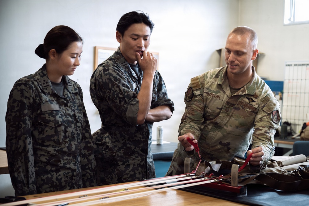 Three military personnel are gathered around a workbench examining equipment. A U.S. Air Force servicemember named MacMillan is demonstrating or explaining items to two individuals wearing what appears to be Republic of Korea (ROK) Air Force digital camouflage uniforms. The setting appears to be a maintenance or training facility.