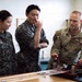 Three military personnel are gathered around a workbench examining equipment. A U.S. Air Force servicemember named MacMillan is demonstrating or explaining items to two individuals wearing what appears to be Republic of Korea (ROK) Air Force digital camouflage uniforms. The setting appears to be a maintenance or training facility.