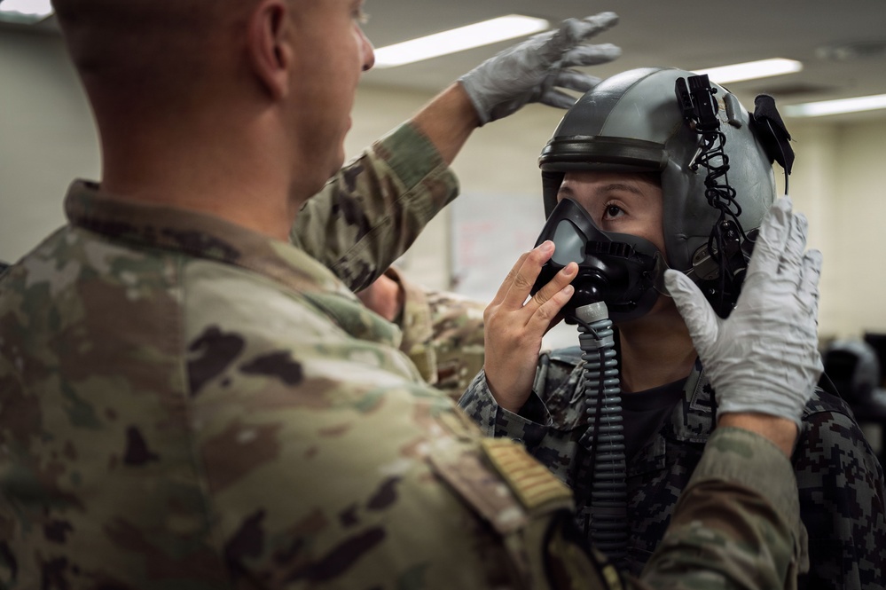 A military personnel in MultiCam/OCP camouflage uniform wearing white latex gloves is fitting or adjusting a flight helmet and oxygen mask on another individual wearing a different camouflage pattern uniform. The scene appears to take place in an indoor facility, likely a life support or equipment fitting room. This appears to be a pre-flight equipment check or fitting procedure.