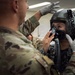 A military personnel in MultiCam/OCP camouflage uniform wearing white latex gloves is fitting or adjusting a flight helmet and oxygen mask on another individual wearing a different camouflage pattern uniform. The scene appears to take place in an indoor facility, likely a life support or equipment fitting room. This appears to be a pre-flight equipment check or fitting procedure.