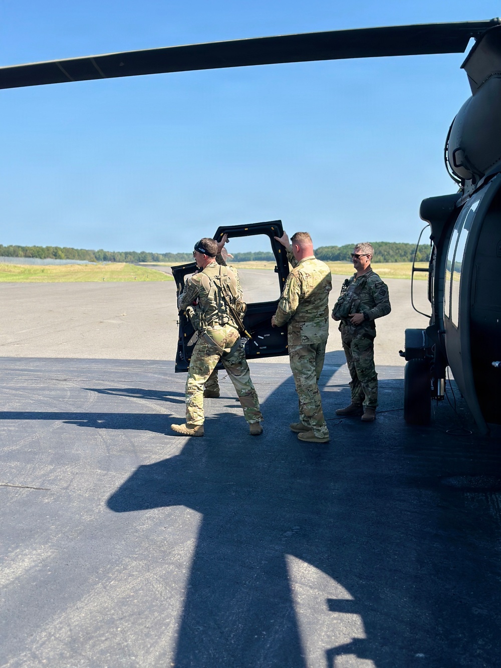 Three military personnel in MultiCam/OCP camouflage uniforms are standing on a tarmac or airfield apron next to a military helicopter, appearing to be preparing for or returning from a mission. One individual is boarding or exiting the aircraft while another holds the door open. The setting appears to be a small airfield with a clear blue sky and tree line visible in the background.