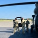 Three military personnel in MultiCam/OCP camouflage uniforms are standing on a tarmac or airfield apron next to a military helicopter, appearing to be preparing for or returning from a mission. One individual is boarding or exiting the aircraft while another holds the door open. The setting appears to be a small airfield with a clear blue sky and tree line visible in the background.