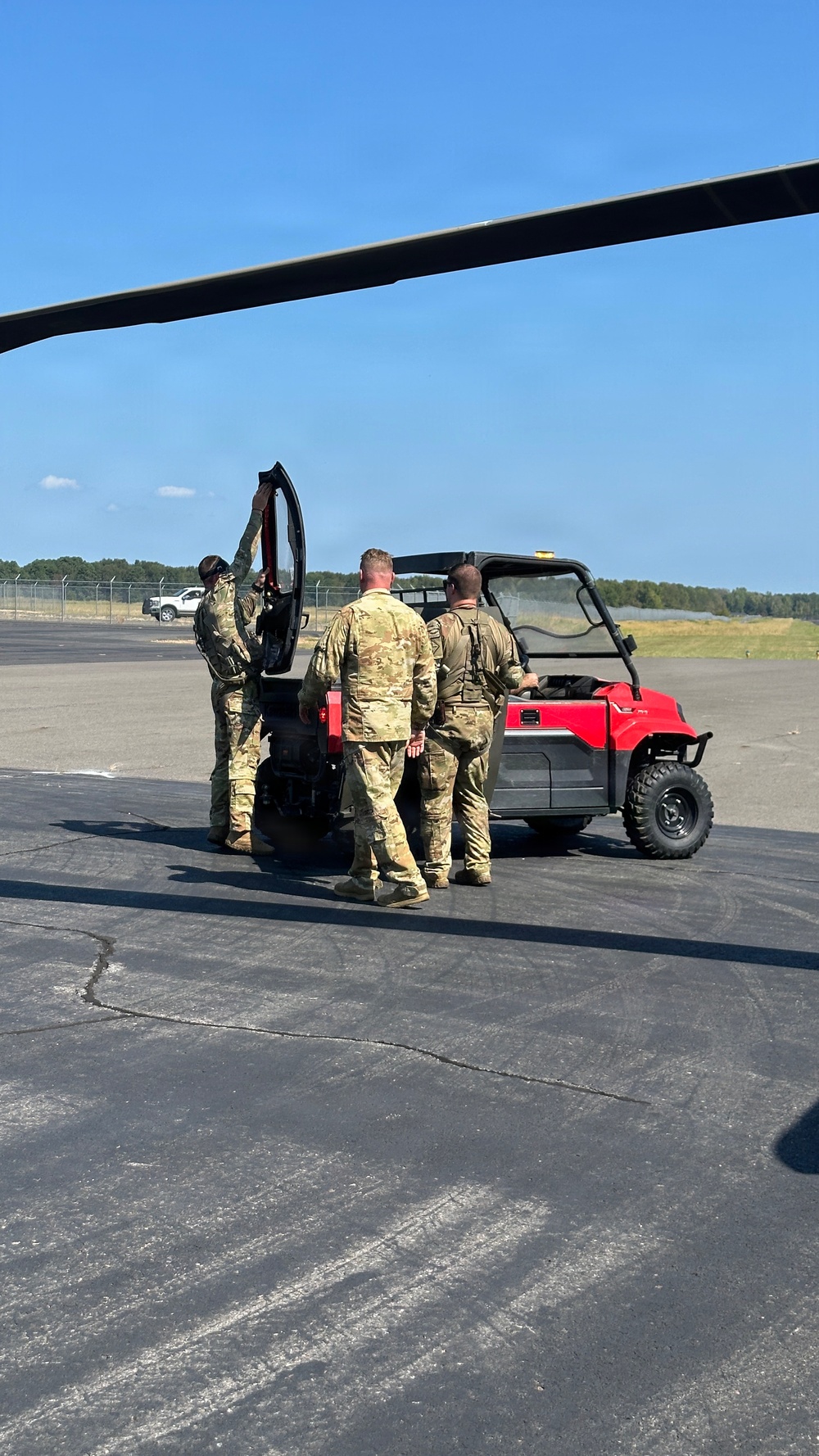 Three military personnel in MultiCam/OCP camouflage uniforms are gathered around a red utility terrain vehicle (UTV) on a tarmac or airfield surface. A large rotor blade is visible in the foreground, indicating proximity to a helicopter. The setting appears to be a military airfield on a clear, sunny day.
