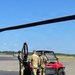 Three military personnel in MultiCam/OCP camouflage uniforms are gathered around a red utility terrain vehicle (UTV) on a tarmac or airfield surface. A large rotor blade is visible in the foreground, indicating proximity to a helicopter. The setting appears to be a military airfield on a clear, sunny day.