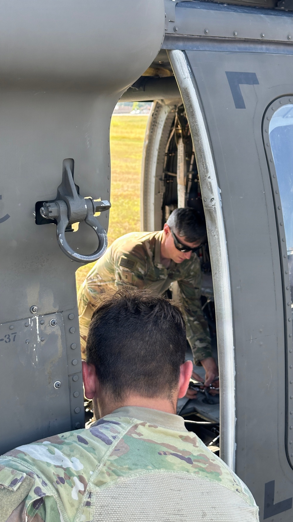 Two military personnel in OCP (Operational Camouflage Pattern) uniforms are performing maintenance or inspection work on a military helicopter. One soldier wearing sunglasses is visible inside the aircraft working on internal components, while a second soldier in the foreground observes or assists. The helicopter appears to be a utility helicopter, likely a UH-60 Black Hawk, based on visible structural features.