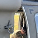 Two military personnel in OCP (Operational Camouflage Pattern) uniforms are performing maintenance or inspection work on a military helicopter. One soldier wearing sunglasses is visible inside the aircraft working on internal components, while a second soldier in the foreground observes or assists. The helicopter appears to be a utility helicopter, likely a UH-60 Black Hawk, based on visible structural features.