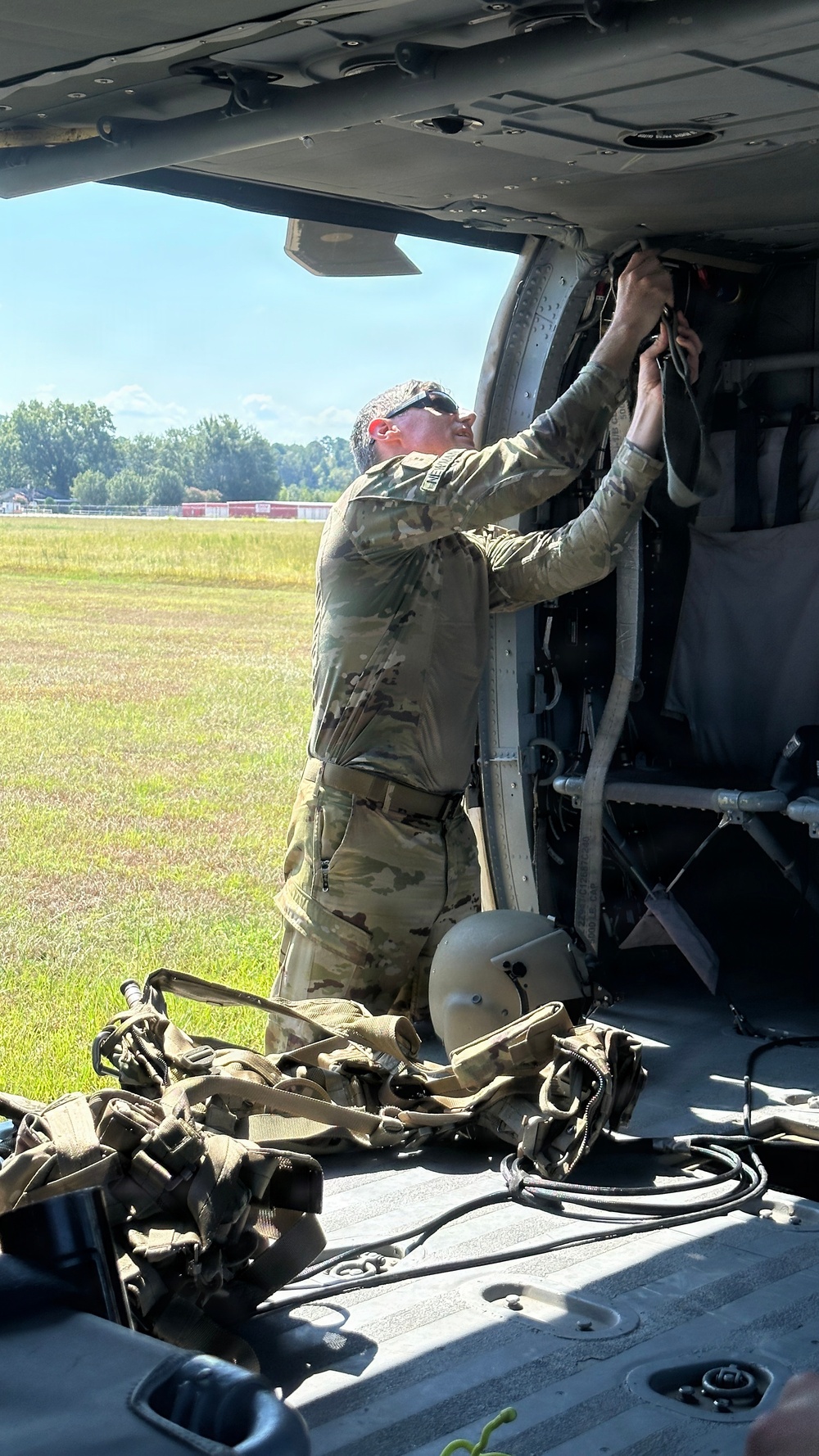 A military service member in OCP (Operational Camouflage Pattern) uniform is performing maintenance or equipment checks on the interior of a military helicopter, likely a UH-60 Black Hawk, while on a grassy airfield. The individual is reaching upward into the aircraft's upper interior panel area, possibly inspecting or adjusting wiring or mechanical components. Various pieces of flight and ground equipment, including a helmet and harness/strap assemblies, are visible on the aircraft floor.