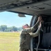 A military service member in OCP (Operational Camouflage Pattern) uniform is performing maintenance or equipment checks on the interior of a military helicopter, likely a UH-60 Black Hawk, while on a grassy airfield. The individual is reaching upward into the aircraft's upper interior panel area, possibly inspecting or adjusting wiring or mechanical components. Various pieces of flight and ground equipment, including a helmet and harness/strap assemblies, are visible on the aircraft floor.