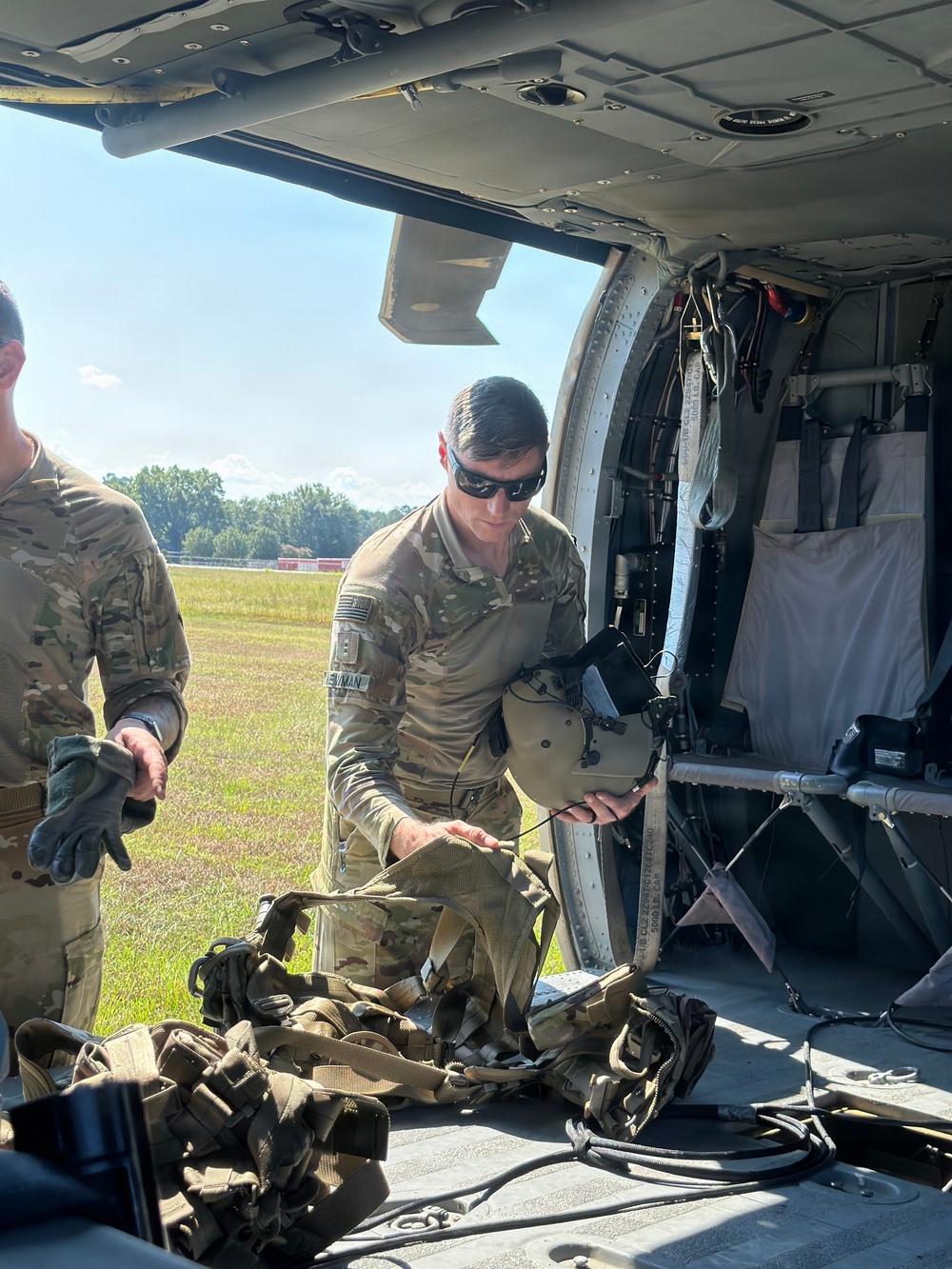 A U.S. Army soldier in OCP (Operational Camouflage Pattern) uniform is conducting pre-flight or post-flight equipment checks at the open door of a military helicopter, likely a UH-60 Black Hawk. The soldier, whose name tape reads 'NEWMAN,' is handling a flight helmet and harness/rigging equipment. A second soldier is partially visible to the left, and the setting appears to be a grassy airfield on a clear day.