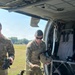 A U.S. Army soldier in OCP (Operational Camouflage Pattern) uniform is conducting pre-flight or post-flight equipment checks at the open door of a military helicopter, likely a UH-60 Black Hawk. The soldier, whose name tape reads 'NEWMAN,' is handling a flight helmet and harness/rigging equipment. A second soldier is partially visible to the left, and the setting appears to be a grassy airfield on a clear day.