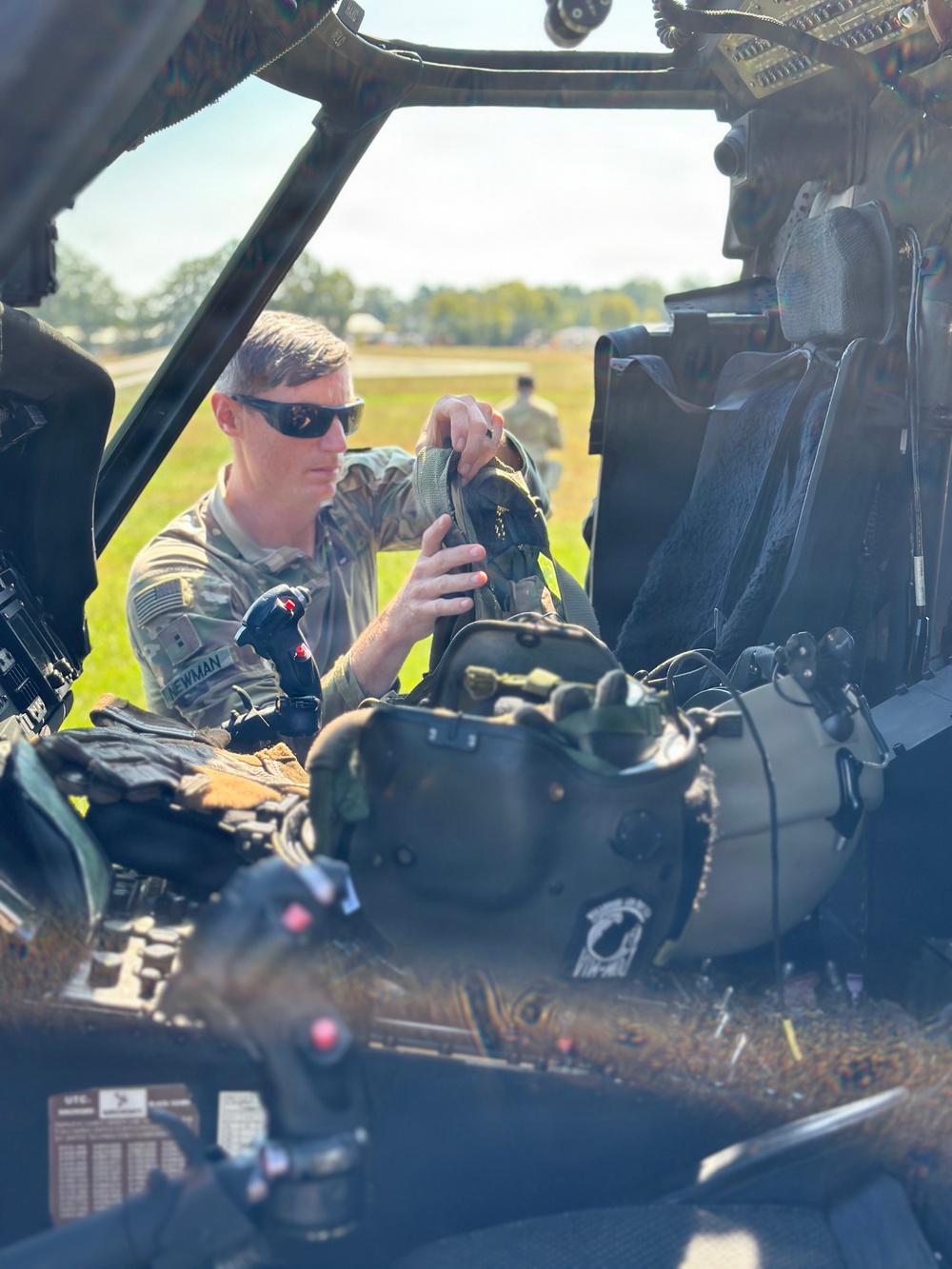 A U.S. Army soldier, identified by nametape as 'NEWMAN', is seated inside what appears to be a military helicopter cockpit, handling flight equipment including a helmet with night vision goggle mounts. The soldier is wearing OCP (Operational Camouflage Pattern) uniform and tactical sunglasses. The setting appears to be a flight line or airfield during daylight hours.