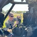 A U.S. Army soldier, identified by nametape as 'NEWMAN', is seated inside what appears to be a military helicopter cockpit, handling flight equipment including a helmet with night vision goggle mounts. The soldier is wearing OCP (Operational Camouflage Pattern) uniform and tactical sunglasses. The setting appears to be a flight line or airfield during daylight hours.