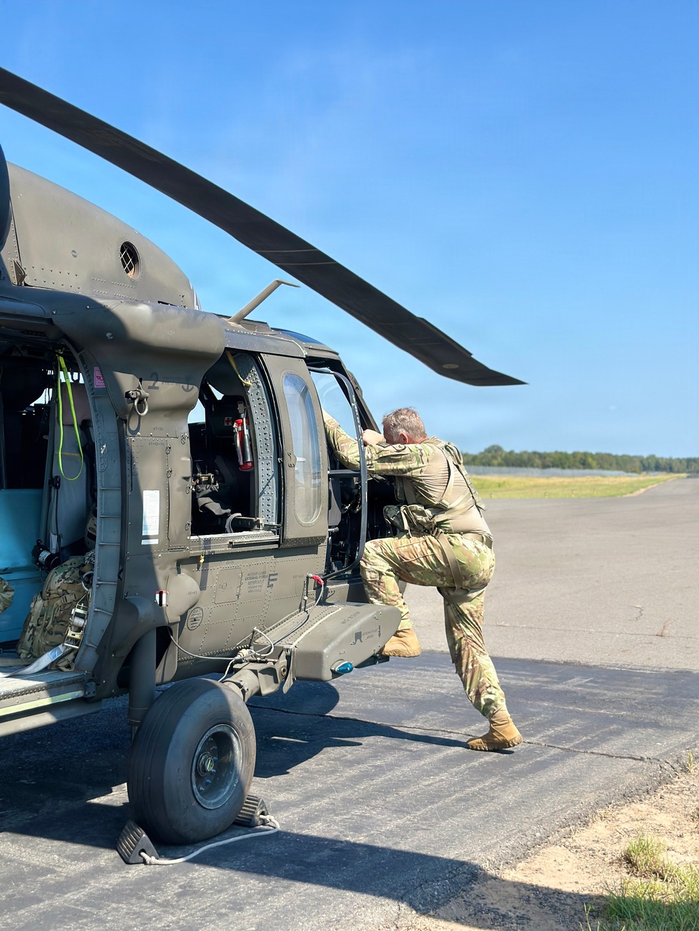 A soldier in MultiCam/OCP camouflage uniform is performing a pre-flight or maintenance inspection on a U.S. Army UH-60 Black Hawk helicopter on a tarmac. The aircraft is stationary with rotor blades visible and a tie-down cable securing the landing gear. The setting appears to be a small airfield or forward operating location on a clear, sunny day.
