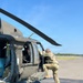A soldier in MultiCam/OCP camouflage uniform is performing a pre-flight or maintenance inspection on a U.S. Army UH-60 Black Hawk helicopter on a tarmac. The aircraft is stationary with rotor blades visible and a tie-down cable securing the landing gear. The setting appears to be a small airfield or forward operating location on a clear, sunny day.