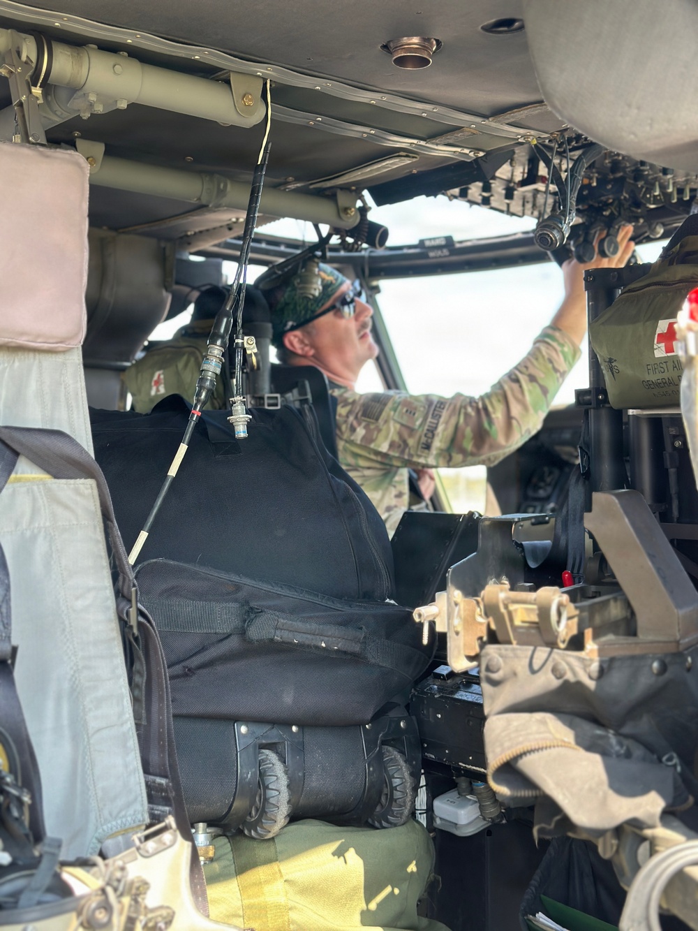 A military personnel member in camouflage uniform is seated inside what appears to be a UH-60 Black Hawk helicopter cockpit, reaching upward to interact with overhead equipment or controls. The individual is wearing a camouflage cap, sunglasses, and a name tape visible on their uniform. A Red Cross first aid kit is visible on the right side, suggesting this may be a medevac or medical evacuation aircraft.
