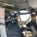 A military personnel member in camouflage uniform is seated inside what appears to be a UH-60 Black Hawk helicopter cockpit, reaching upward to interact with overhead equipment or controls. The individual is wearing a camouflage cap, sunglasses, and a name tape visible on their uniform. A Red Cross first aid kit is visible on the right side, suggesting this may be a medevac or medical evacuation aircraft.