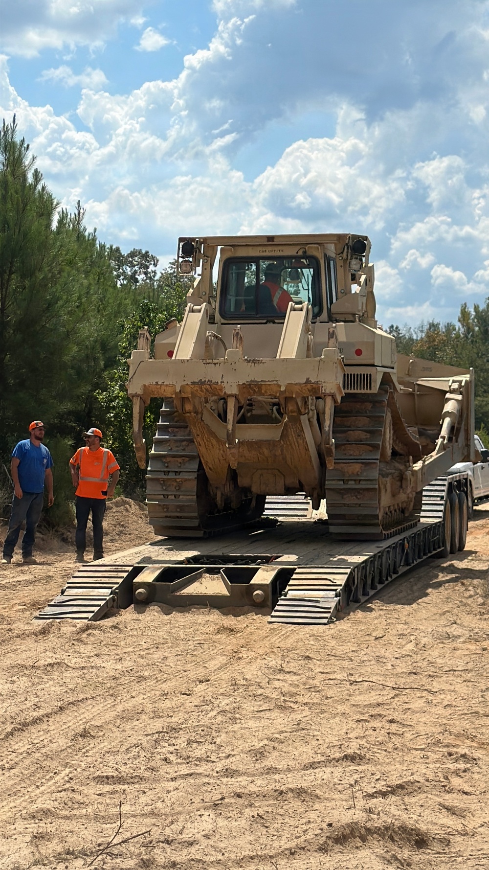 A large tracked bulldozer, painted in a tan/beige color, is being unloaded from a lowboy trailer on a sandy dirt surface. Two workers are standing nearby observing the operation, one wearing an orange high-visibility vest and the other in a blue shirt, both with hard hats. The scene is set in a wooded area under a partly cloudy sky.