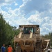 A large tracked bulldozer, painted in a tan/beige color, is being unloaded from a lowboy trailer on a sandy dirt surface. Two workers are standing nearby observing the operation, one wearing an orange high-visibility vest and the other in a blue shirt, both with hard hats. The scene is set in a wooded area under a partly cloudy sky.