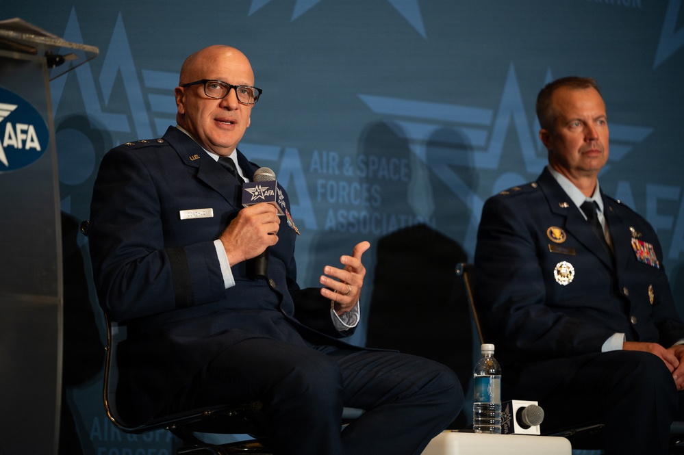 Two military officers in US Air Force dress blue uniforms are seated on a panel stage at an Air &amp; Space Forces Association (AFA) event. The officer on the left is speaking into an AFA-branded microphone while gesturing with his hand. The backdrop prominently displays the AFA logo and 'Air &amp; Space Forces Association' branding.