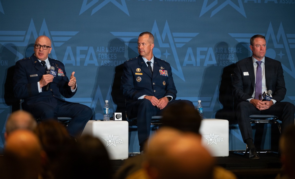 Three individuals are seated on a panel stage at an AFA (Air &amp; Space Forces Association) event. Two are wearing United States Air Force dress uniforms with visible medals and insignia, while the third is in civilian business attire. The leftmost uniformed individual is speaking into a microphone while the others listen.
