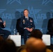 Three individuals are seated on a panel stage at an AFA (Air &amp; Space Forces Association) event. Two are wearing United States Air Force dress uniforms with visible medals and insignia, while the third is in civilian business attire. The leftmost uniformed individual is speaking into a microphone while the others listen.
