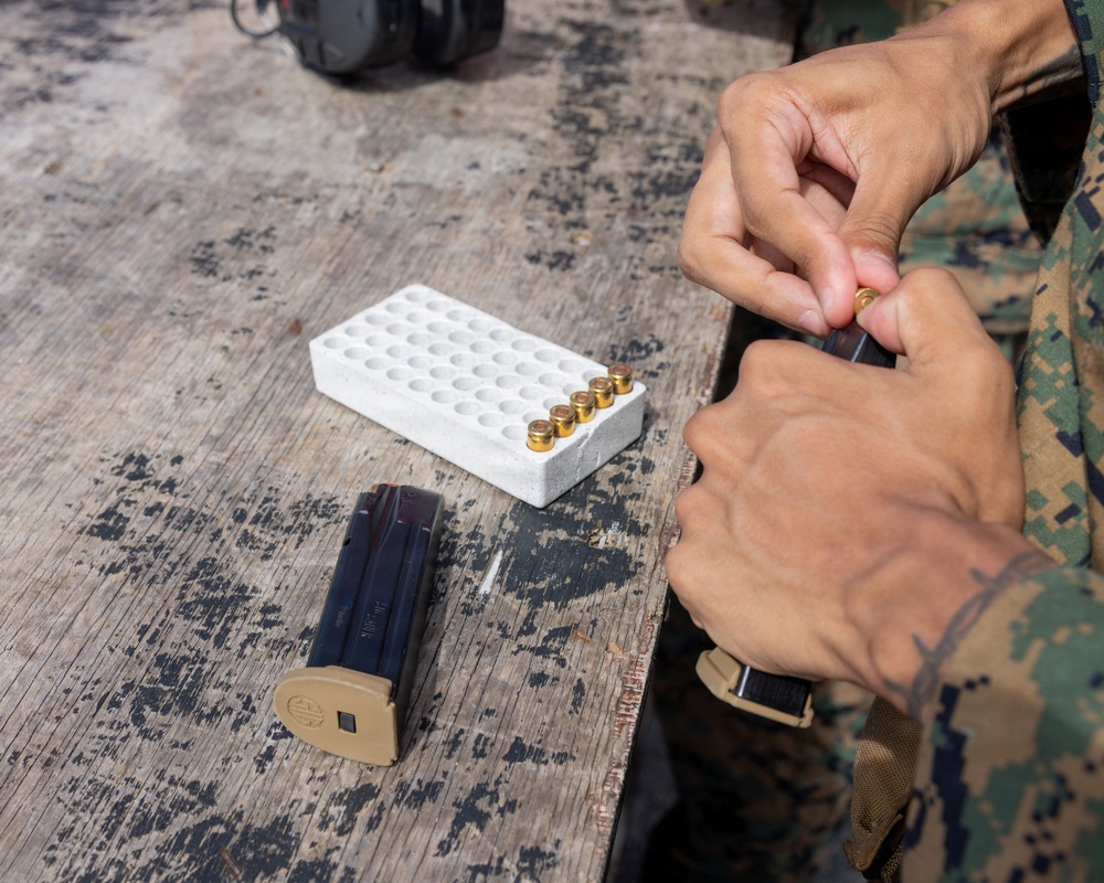 A person wearing U.S. Marine Corps MARPAT digital camouflage uniform is loading ammunition into a pistol magazine at an outdoor shooting range or training area. A white foam ammunition tray with several brass cartridges is visible on a weathered wooden table, along with a second magazine. The setting appears to be a military firearms qualification or training exercise.