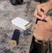 A person wearing U.S. Marine Corps MARPAT digital camouflage uniform is loading ammunition into a pistol magazine at an outdoor shooting range or training area. A white foam ammunition tray with several brass cartridges is visible on a weathered wooden table, along with a second magazine. The setting appears to be a military firearms qualification or training exercise.