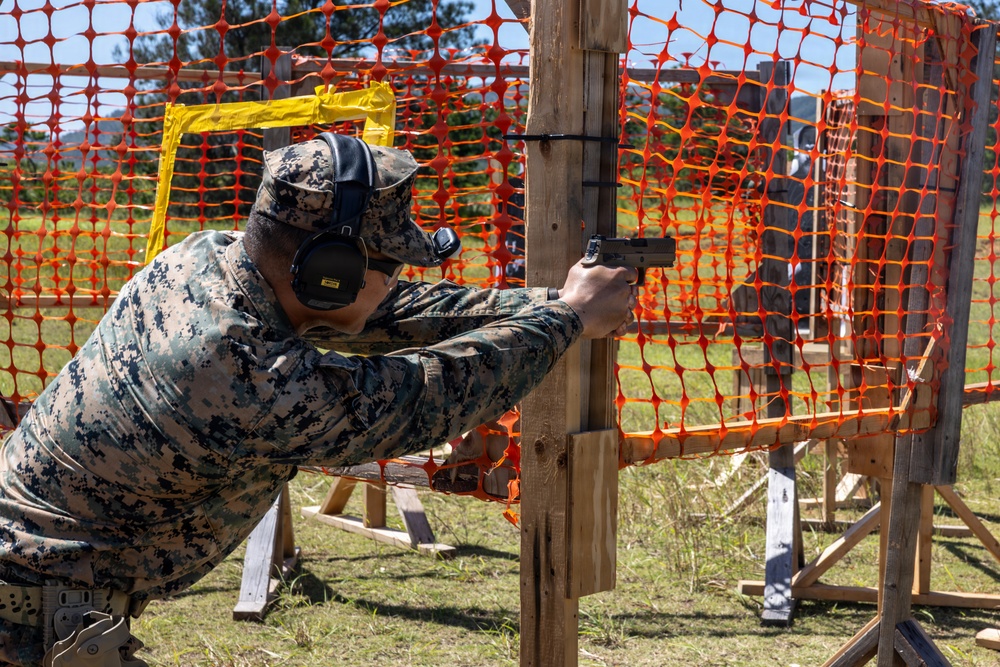 A military service member in digital camouflage uniform is engaged in a pistol shooting exercise at an outdoor range. The shooter is using a wooden post as cover while aiming a semi-automatic pistol downrange. The setting appears to be a structured tactical shooting course with orange safety fencing and wooden frame obstacles.