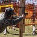 A military service member in digital camouflage uniform is engaged in a pistol shooting exercise at an outdoor range. The shooter is using a wooden post as cover while aiming a semi-automatic pistol downrange. The setting appears to be a structured tactical shooting course with orange safety fencing and wooden frame obstacles.