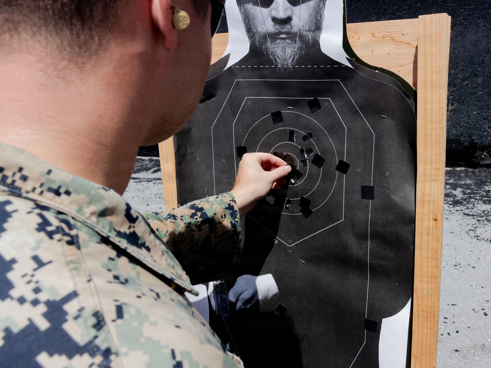 A military service member in digital camouflage uniform is examining a paper shooting target at a firing range. The target shows numerous bullet holes concentrated around the center scoring rings, indicating marksmanship evaluation. The person appears to be scoring or patching the target after a qualification exercise.