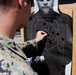 A military service member in digital camouflage uniform is examining a paper shooting target at a firing range. The target shows numerous bullet holes concentrated around the center scoring rings, indicating marksmanship evaluation. The person appears to be scoring or patching the target after a qualification exercise.