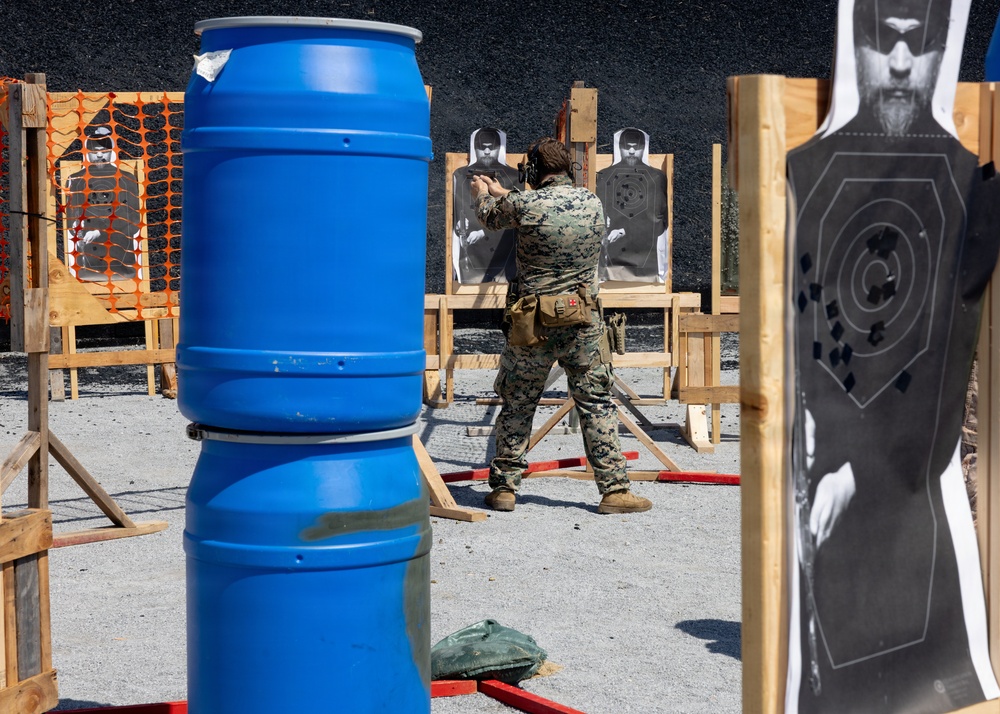 A military or law enforcement personnel in camouflage uniform is engaged in a shooting range exercise, aiming a firearm at paper targets mounted on wooden frames. The range features multiple human-silhouette targets showing bullet impact marks, blue plastic barrels used as cover obstacles, and orange safety fencing in the background. The setting appears to be an outdoor tactical shooting course designed to simulate real-world engagement scenarios.