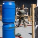 A military or law enforcement personnel in camouflage uniform is engaged in a shooting range exercise, aiming a firearm at paper targets mounted on wooden frames. The range features multiple human-silhouette targets showing bullet impact marks, blue plastic barrels used as cover obstacles, and orange safety fencing in the background. The setting appears to be an outdoor tactical shooting course designed to simulate real-world engagement scenarios.