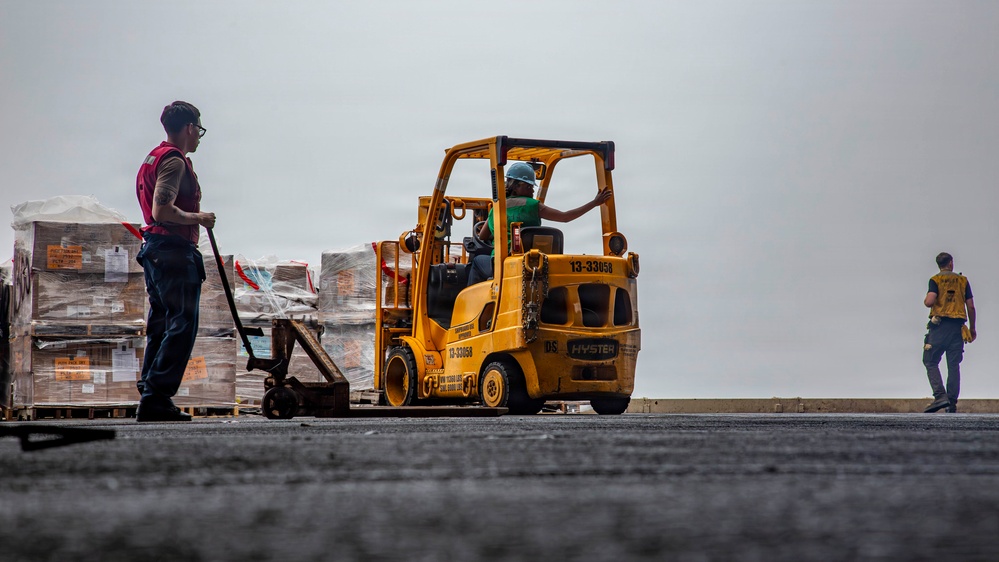 A logistics or cargo handling operation is underway on what appears to be the deck of a naval vessel or large industrial platform. A yellow Hyster forklift (marked '13-33058') is being operated by a worker wearing a blue hard hat and green shirt, while a second worker in a red vest and glasses stands nearby monitoring palletized cargo. A third worker in a yellow vest is visible walking in the background.
