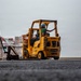 A logistics or cargo handling operation is underway on what appears to be the deck of a naval vessel or large industrial platform. A yellow Hyster forklift (marked '13-33058') is being operated by a worker wearing a blue hard hat and green shirt, while a second worker in a red vest and glasses stands nearby monitoring palletized cargo. A third worker in a yellow vest is visible walking in the background.