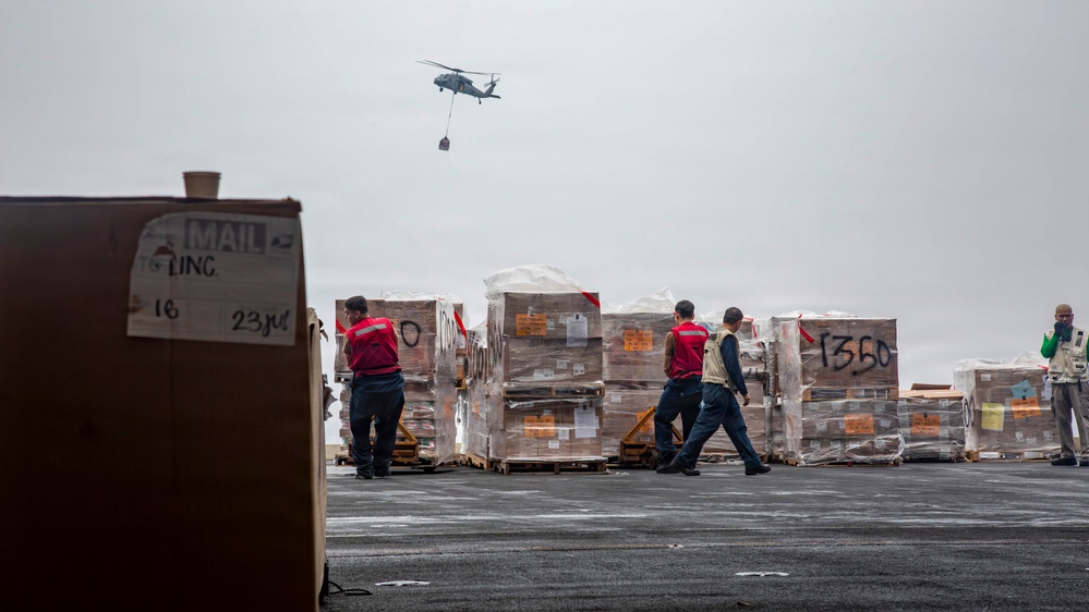 A military or naval logistics operation is underway on what appears to be the deck of an aircraft carrier or large naval vessel. Personnel in red safety vests are managing palletized cargo while a helicopter performs a vertical replenishment (VERTREP) operation overhead, carrying a slung load. Multiple pallets of supplies, including what appears to be mail and other cargo, are staged on the deck.