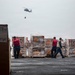 A military or naval logistics operation is underway on what appears to be the deck of an aircraft carrier or large naval vessel. Personnel in red safety vests are managing palletized cargo while a helicopter performs a vertical replenishment (VERTREP) operation overhead, carrying a slung load. Multiple pallets of supplies, including what appears to be mail and other cargo, are staged on the deck.