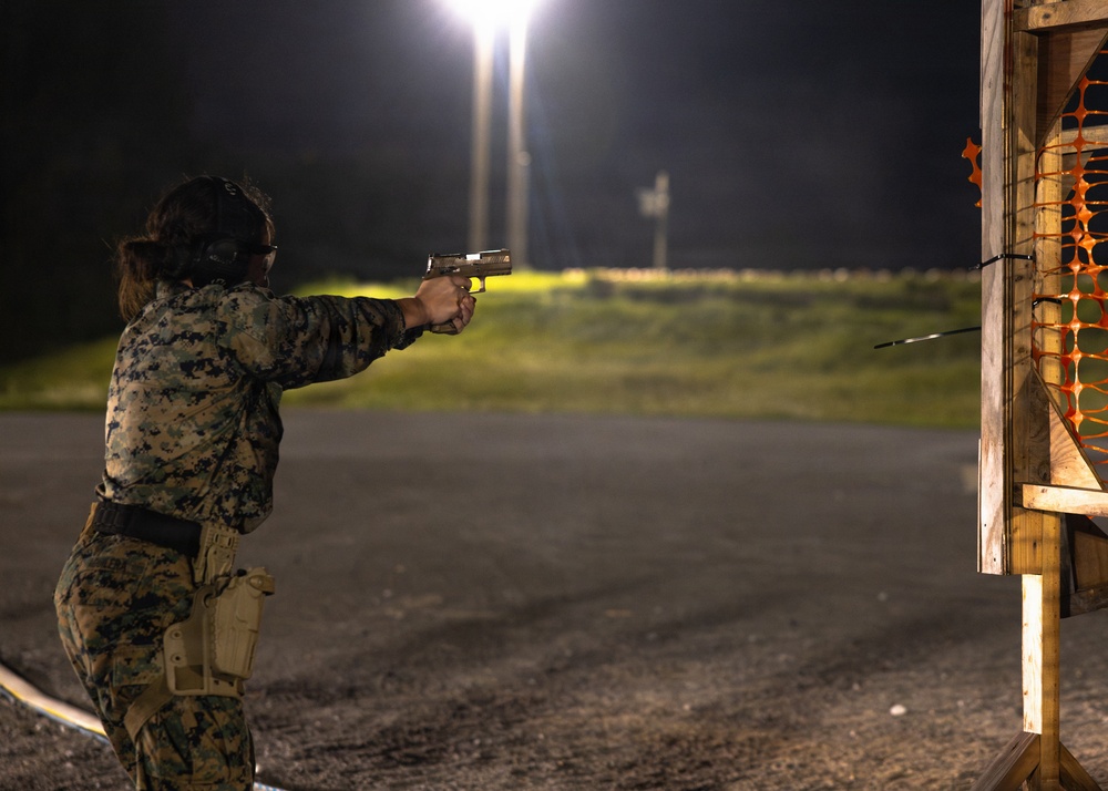 A military service member in Marine Corps MARPAT camouflage uniform is conducting a nighttime pistol shooting exercise at an outdoor range. The shooter is in a two-handed firing stance, aiming a semi-automatic pistol toward a wooden target structure to the right. The scene is illuminated by overhead range lighting against a dark night sky.