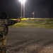 A military service member in Marine Corps MARPAT camouflage uniform is conducting a nighttime pistol shooting exercise at an outdoor range. The shooter is in a two-handed firing stance, aiming a semi-automatic pistol toward a wooden target structure to the right. The scene is illuminated by overhead range lighting against a dark night sky.