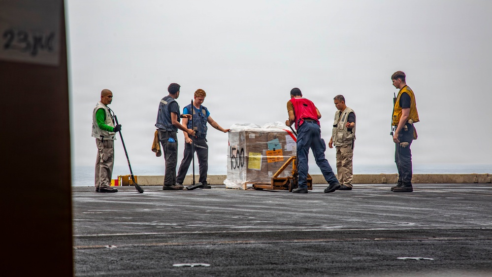 Six personnel are handling a wrapped pallet of cargo on what appears to be the flight deck of a naval vessel. The pallet is marked with the number '1500' and has orange and yellow labels attached. The scene takes place under overcast skies with the ocean visible in the background.