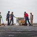Six personnel are handling a wrapped pallet of cargo on what appears to be the flight deck of a naval vessel. The pallet is marked with the number '1500' and has orange and yellow labels attached. The scene takes place under overcast skies with the ocean visible in the background.