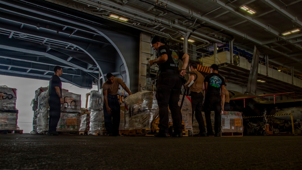 Several personnel are working inside what appears to be the hangar bay of a naval vessel, handling palletized cargo wrapped in plastic. The large open bay door in the background reveals an overcast exterior, and a helicopter or aircraft is partially visible to the right. The scene depicts a logistics or resupply operation taking place aboard a warship.