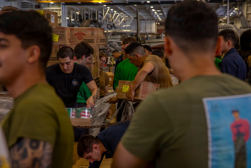 A group of military personnel, likely U.S. Navy sailors and/or Marines, are working together to unload and sort supplies in what appears to be the hangar bay of a naval vessel. They are handling cardboard boxes and palletized cargo, suggesting a replenishment-at-sea or stores loading operation. The setting is an enclosed, industrial shipboard space with overhead equipment and lighting visible.
