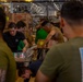 A group of military personnel, likely U.S. Navy sailors and/or Marines, are working together to unload and sort supplies in what appears to be the hangar bay of a naval vessel. They are handling cardboard boxes and palletized cargo, suggesting a replenishment-at-sea or stores loading operation. The setting is an enclosed, industrial shipboard space with overhead equipment and lighting visible.