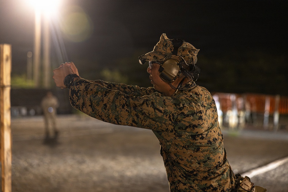 A military service member in digital camouflage uniform is firing a pistol at a nighttime shooting range. The shooter is using a two-handed grip stance with arms extended, wearing hearing protection and eye protection. The scene is illuminated by artificial range lighting against a dark background.